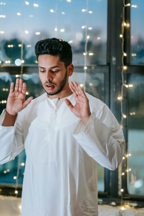 A Muslim person standing upright in the Qiyam position, the initial posture of Salah, with hands raised to the ears.